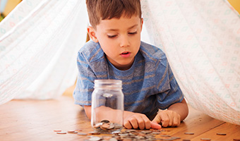 Little boy adding coins to jar full of money laying down under tent made from bed sheet.